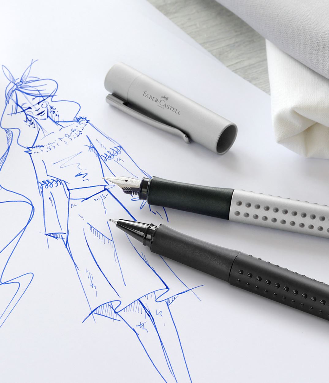 Grey grip fountain pen and black grip finewriter lying on a paper with a blue fashion drawing.
